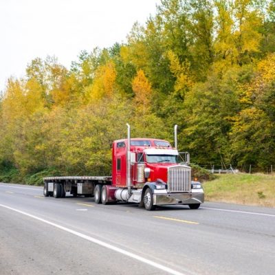 Red American icon big rig classic bonnet powerful long haul semi truck with vertical tailpipes and empty flat bed semi trailer running on the autumn road with green and yellow trees to loading point